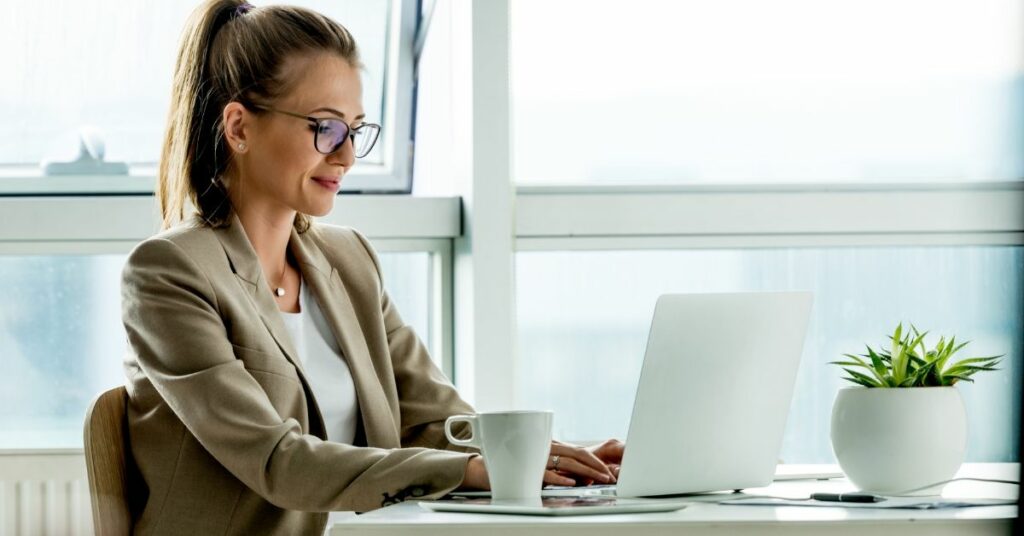 Young white woman with glasses sitting at her desk working on her laptop with a cup of coffee.